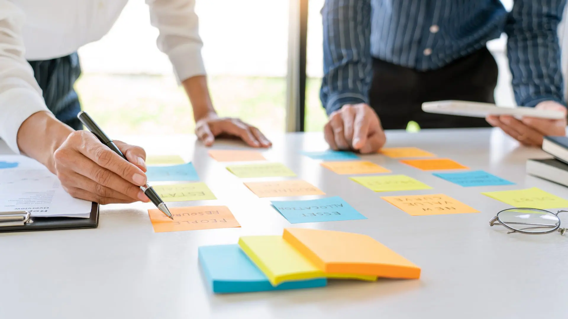 workplace close-up of a white table with colorful post-it notes and pairs of hands, one pointing, one with a pen, representing planning/strategizing