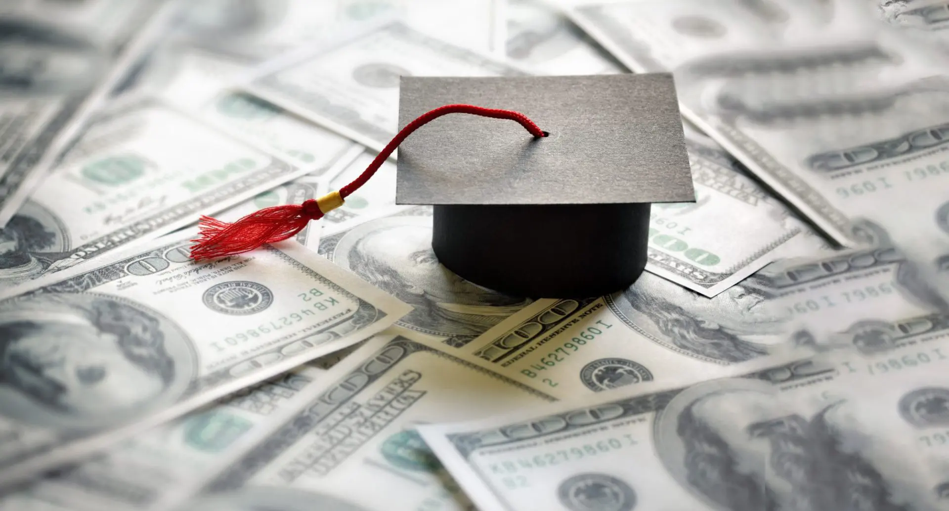 Graduation mortarboard with red tassel atop a spread of dollar bills, representing student loan debt