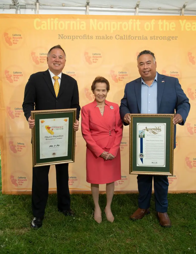 Two men in suits with a woman in a red dress in a middle against a Nonprofit of the Year banner