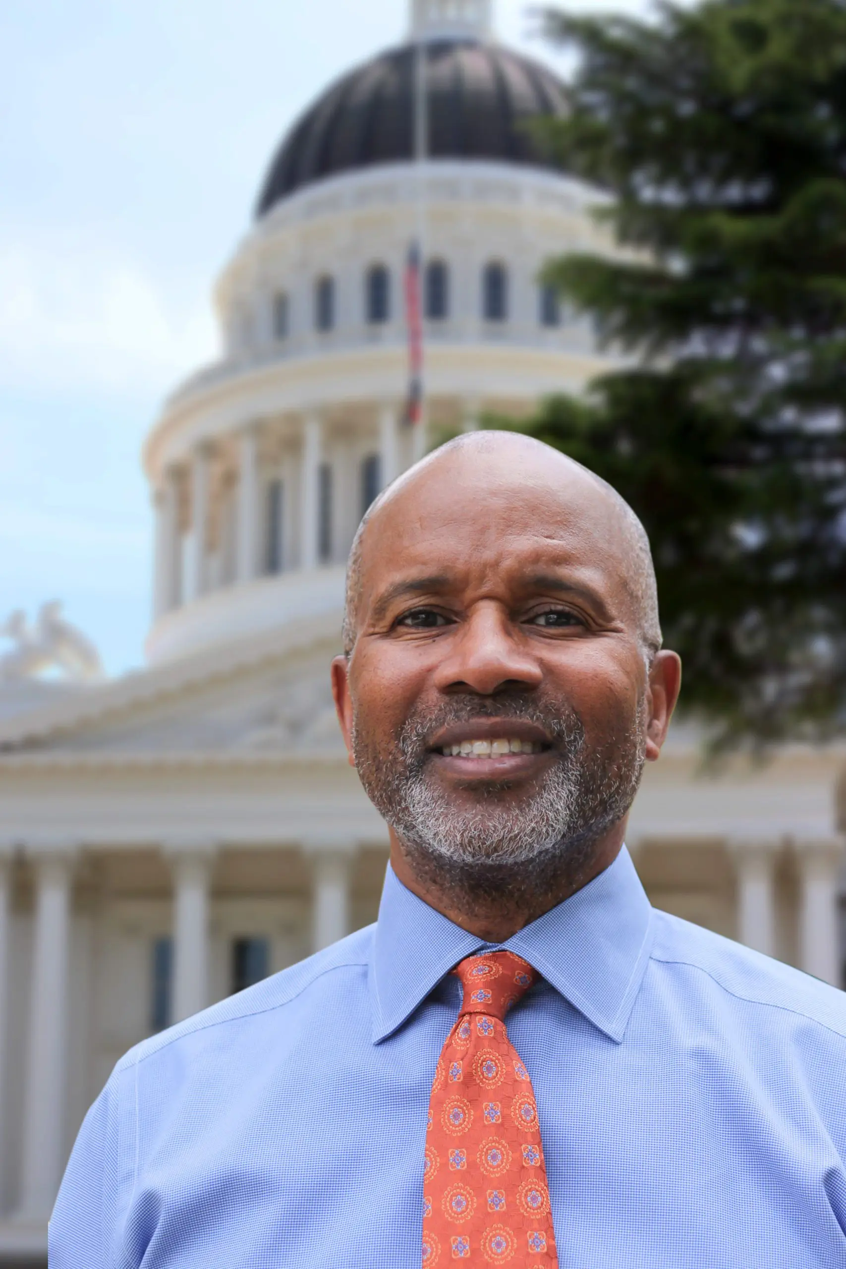 Arnie Sowell, headshot, a middle age African American man wwith a trim grey beard and wearing a light blue shirt and red tie. CA state Capitol building in background.