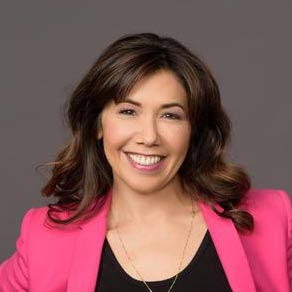 Jody Hicks, headshot of a woman with wavy shoulder length hair, wearing a black tee and hot pink jacket. Smiling.