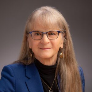 Headshot, Pamela David, a mature white woman, with blonde long hai, glasses, wearing a black turtleneck and dark blue jacket